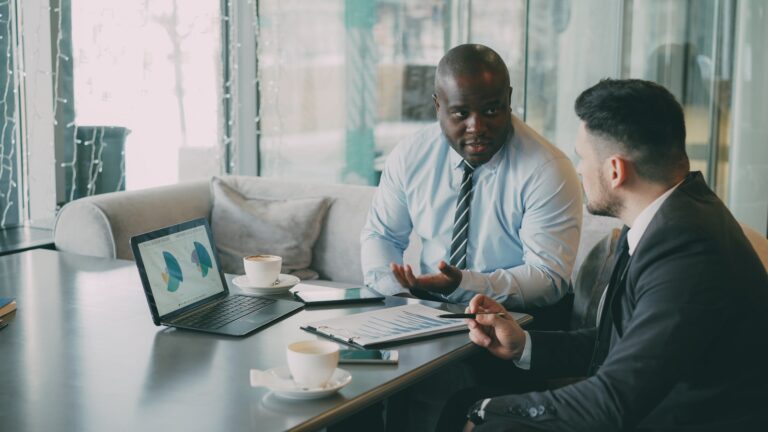 Two businessmen reviewing sales charts on a laptop and printed report during a meeting