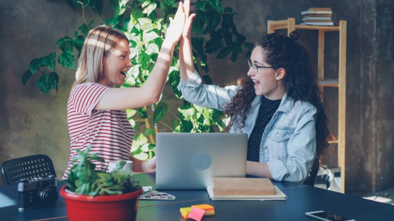 Two coworkers celebrating a business win with a high five at their desk